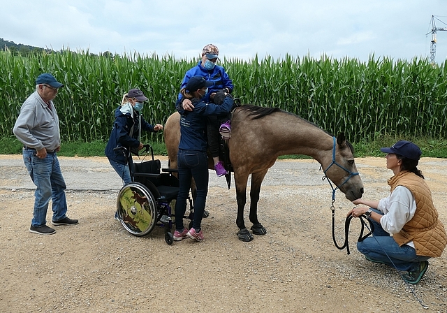 Entspannt und glücklich wird die halbseitig gelähmte Patientin sicher vom Hippotherapiepferd gehoben und in den mit Pferden verzierten Rollstuhl gesetzt.